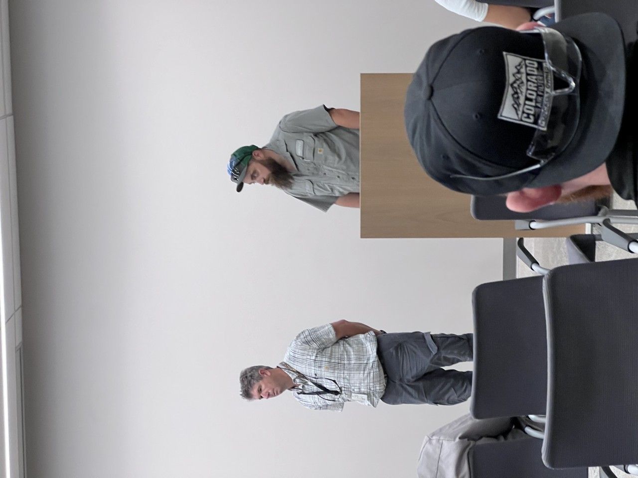 Three men in a room; one at a desk, two in chairs. The man at the desk is looking down, others are looking away.