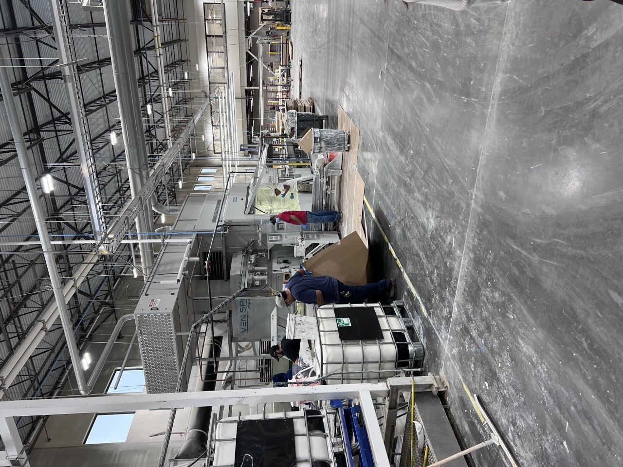 Workers in a factory setting, near industrial machinery and white tanks, on a concrete floor.