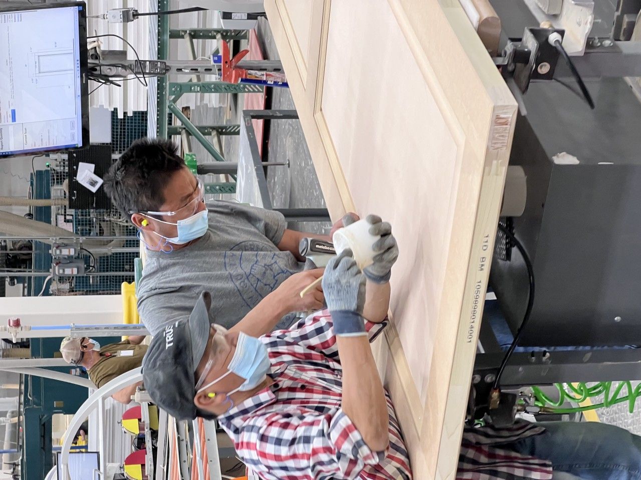 Two workers wearing masks apply glue to a large wooden door in a factory setting.