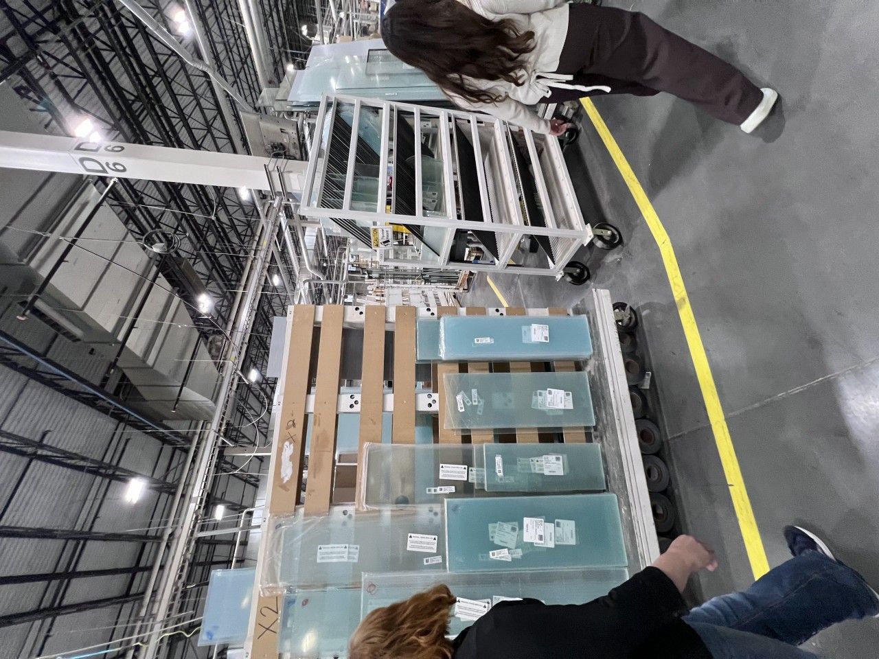 People near a pallet of packaged glass in a warehouse. Overhead crane and yellow line are visible.