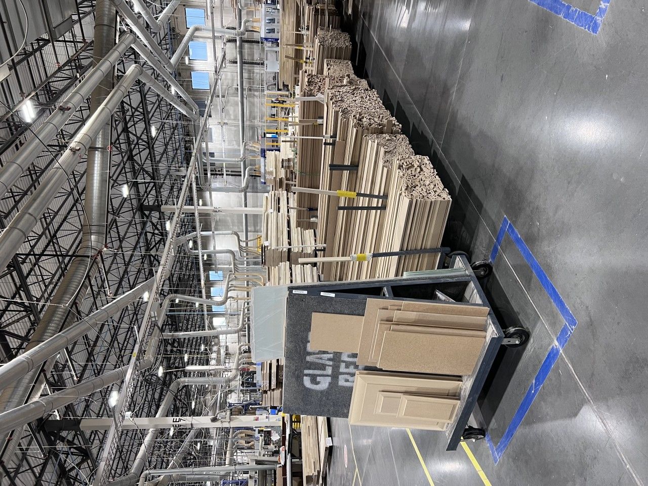 Warehouse interior with stacks of wooden planks, cardboard boxes on a pallet jack, and overhead metal structures.