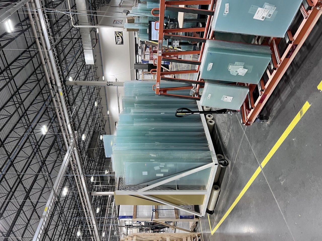 Stacks of glass sheets in a warehouse setting, with industrial lighting and a yellow line on the floor.