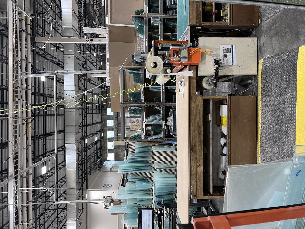 Inside a factory, stacks of glass panels near machinery, with overhead lights and girders.