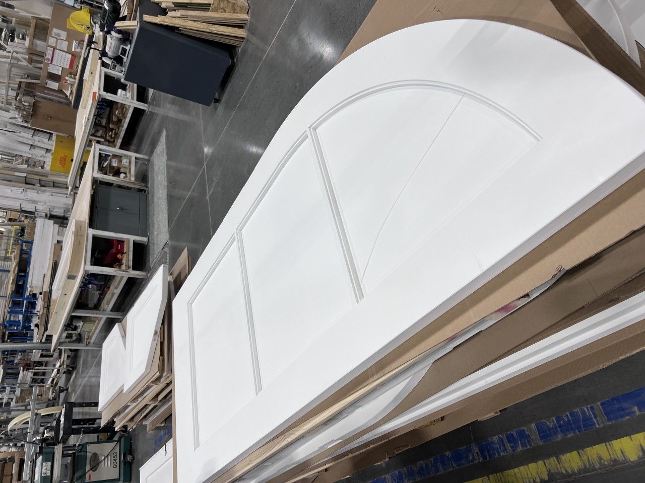 White arched door panel with etched design, sitting on a stack of cardboard and wood materials in a warehouse setting.