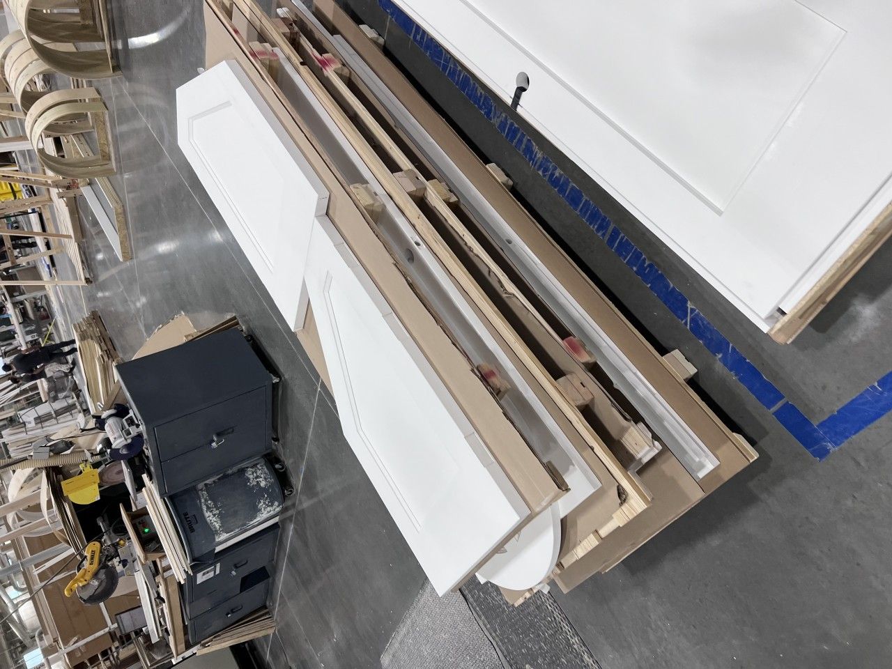 Overhead view of stacked white cabinet doors and long pieces of wood on a factory floor.
