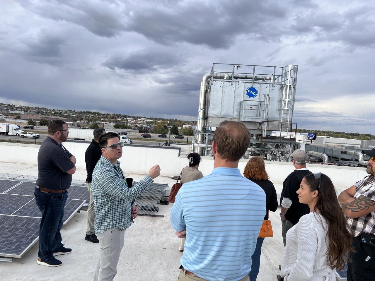 Group of people on a rooftop, looking at equipment. One man gestures with his hand. Cloudy sky overhead.