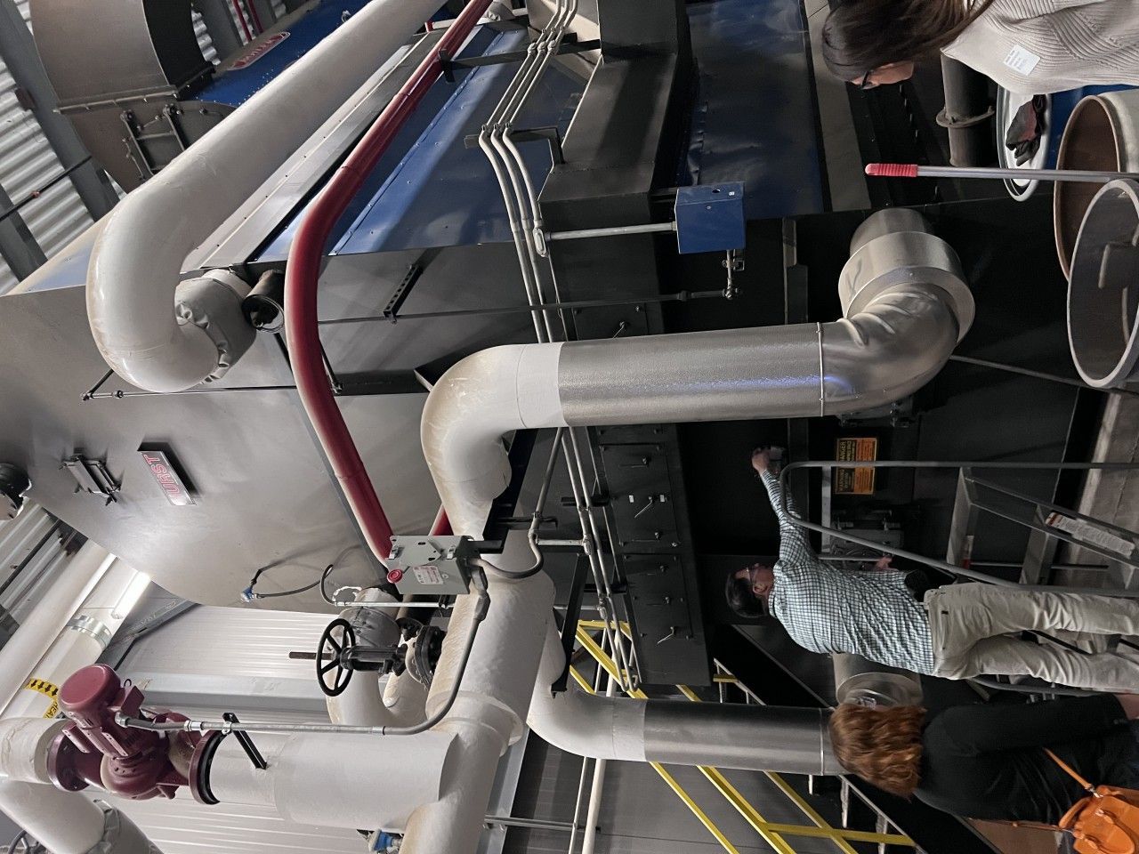 People observing industrial machinery with insulated pipes in a large building.