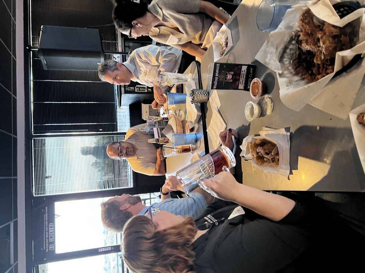 People eating at a restaurant table; food, drinks, overhead light, and a window are visible.