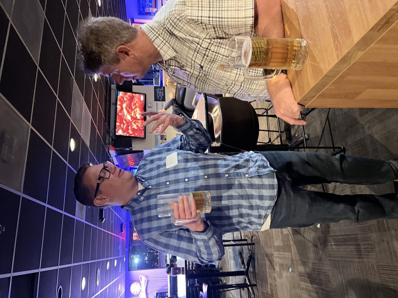 Two men at a bar, holding drinks, one in a blue plaid shirt, the other in a light-colored plaid shirt, conversing.