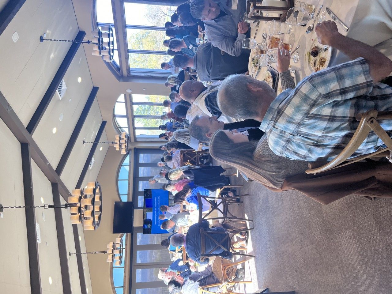 People seated at a long table in a dining room, eating and conversing. Bright interior with large windows.