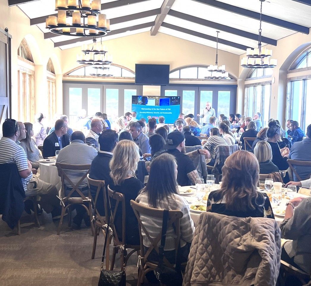 Large gathering in a spacious room. People seated at tables, a speaker at the front, and a presentation on a screen.