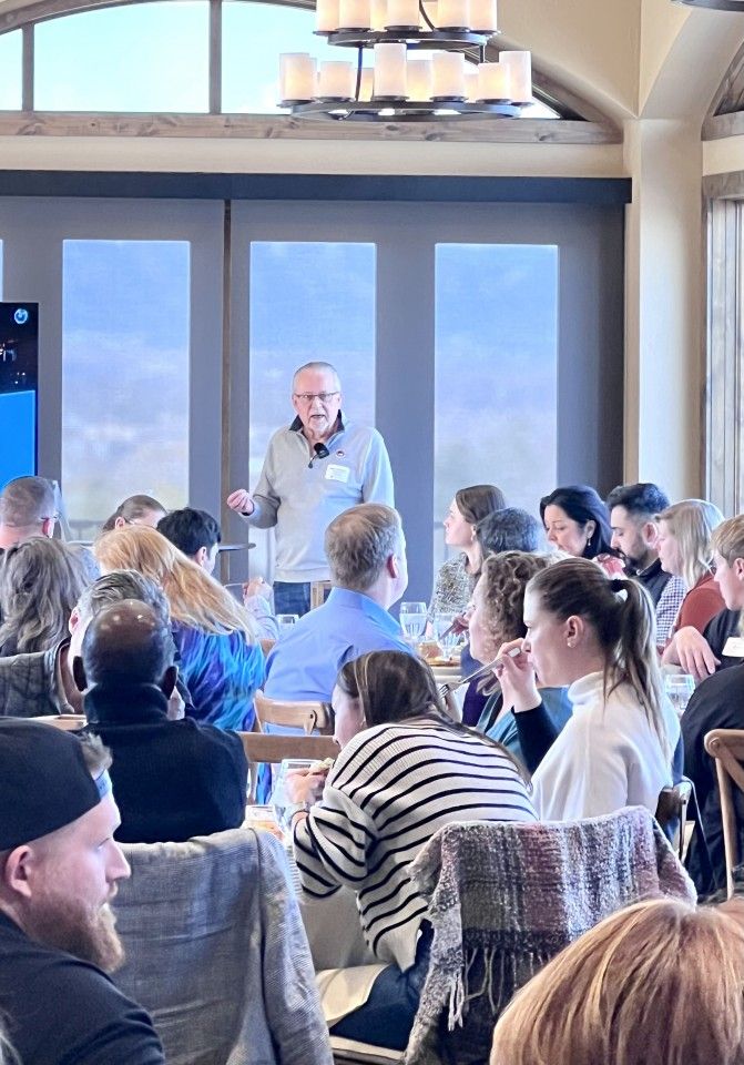 A man speaking to a seated audience in a bright room with large windows.