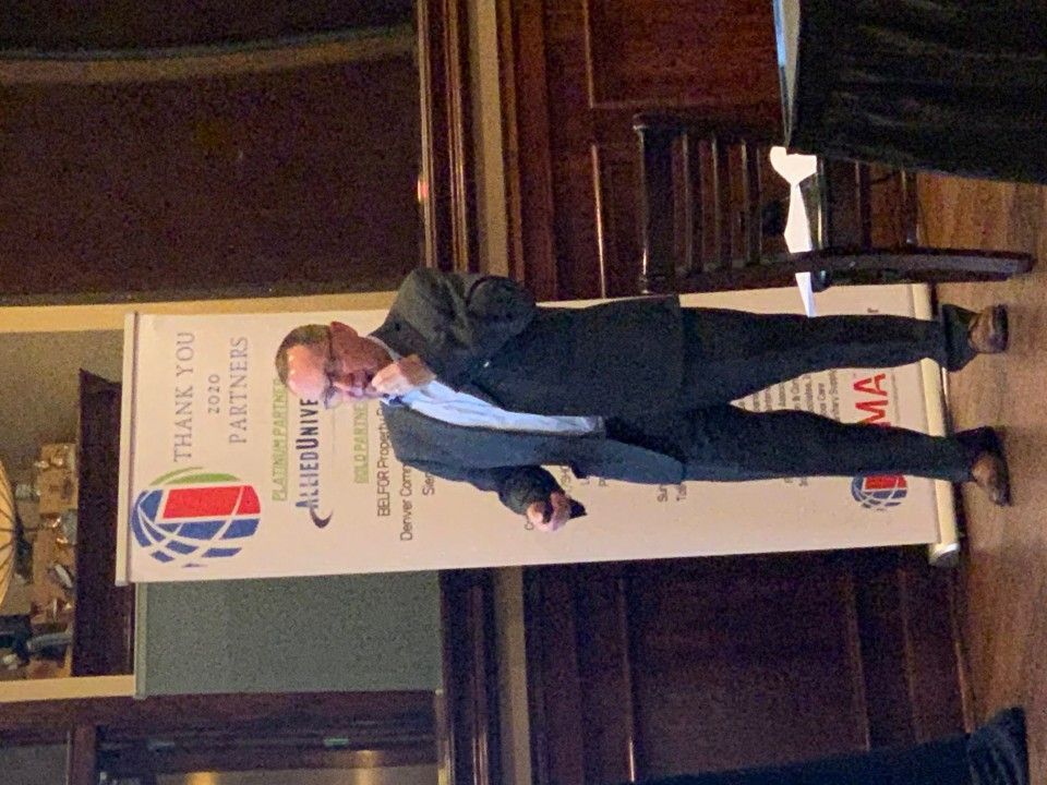 Man in suit speaking at event, next to banner that reads 