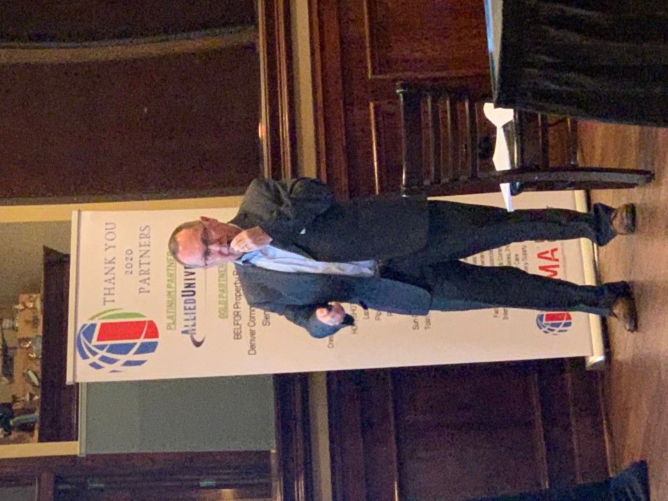Man in suit gestures next to a banner thanking sponsors, in a wood-paneled room.