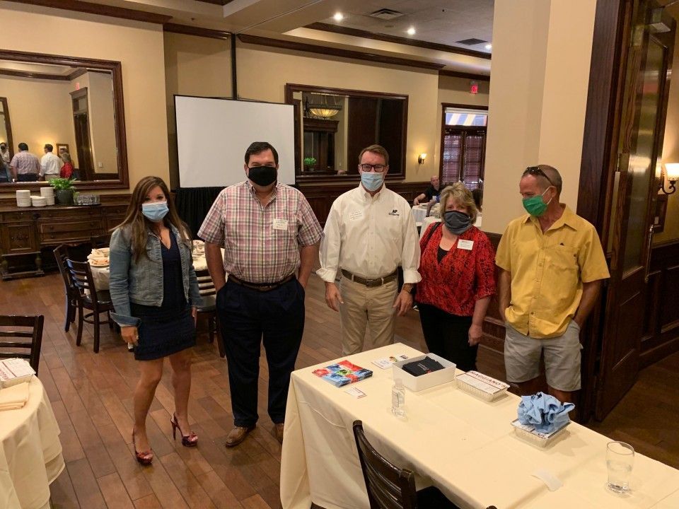 Five masked people stand near a table in a restaurant. White screen and mirror visible.