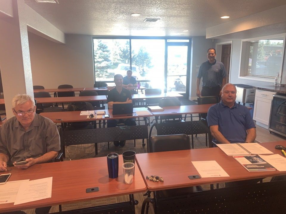 People seated at tables in a classroom setting, a few standing, windows in background.