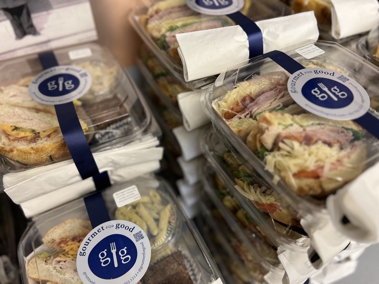 Sandwiches in clear plastic containers, stacked, with blue ribbon and logo sticker.