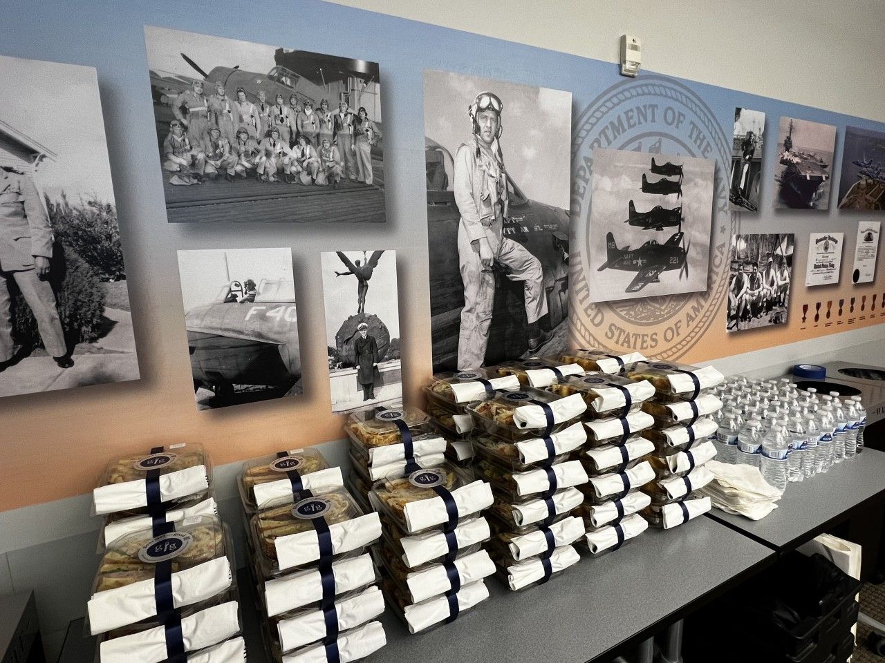 Stacks of packaged food and water bottles on a table in front of historical photos on a wall.