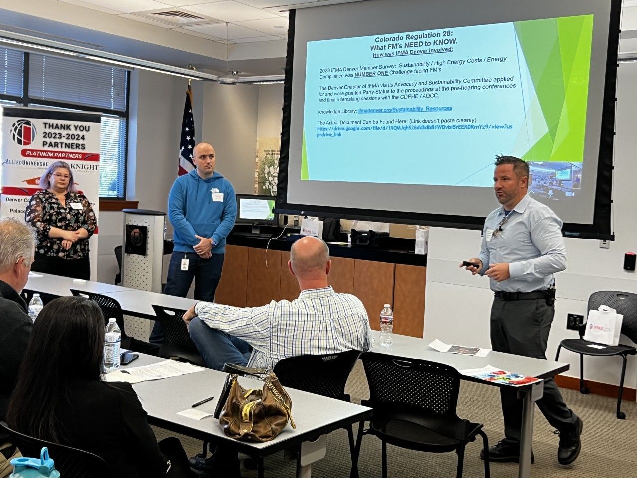 A presentation in a conference room with three speakers, a screen, and an audience.