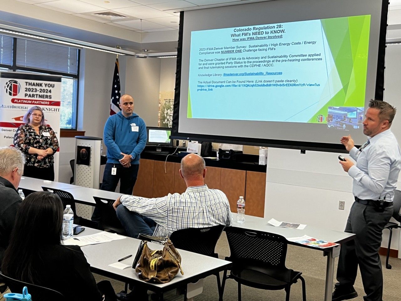 People in a presentation. A man points at a projector screen with text. Others watch, some seated at tables.