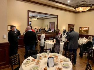People in a well-lit dining room; tables set with food, and a large mirror reflects the room.
