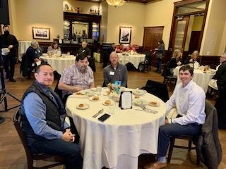 Four people seated at a round table in a restaurant; other diners visible in the background.