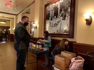 Man in mask buys treats from two girls in a lobby, snacks on table, old photo on wall.