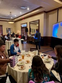 Man speaking in a room. People sit at round tables, some eating. A screen shows a blue graphic.