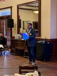 Man speaking into microphone, holding papers, in a dining room with a large mirror and speaker.