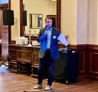 Man in blazer speaking into microphone at an event, holding a paper, with a dark wooden cabinet in the background.