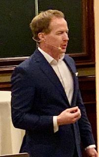 Man in a blue suit speaking, white shirt. He is wearing a lavalier microphone, indoors, looking to his right.