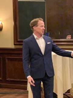 Man in a blue suit speaks, gesturing with his right hand, standing near a table in a room.