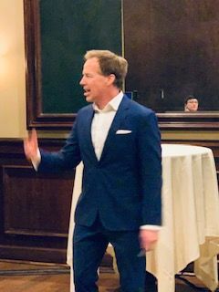 Man in a blue suit gestures while speaking, standing near a table with a white cloth, in a room.