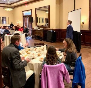 A man speaks at a presentation in a dining room. Attendees sit at round tables.