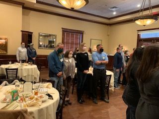 People standing in a restaurant, some wearing masks, observing an event. Tables are set with white tablecloths.