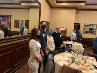 People standing in a restaurant, some wearing masks, by tables set for a meal.