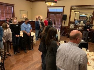 People standing indoors, some wearing masks, facing a speaker. Tables set, likely an event.