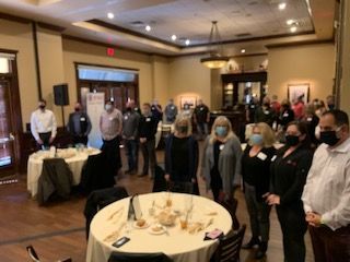 Group of people standing indoors, some wearing masks, by round tables with food and drinks.