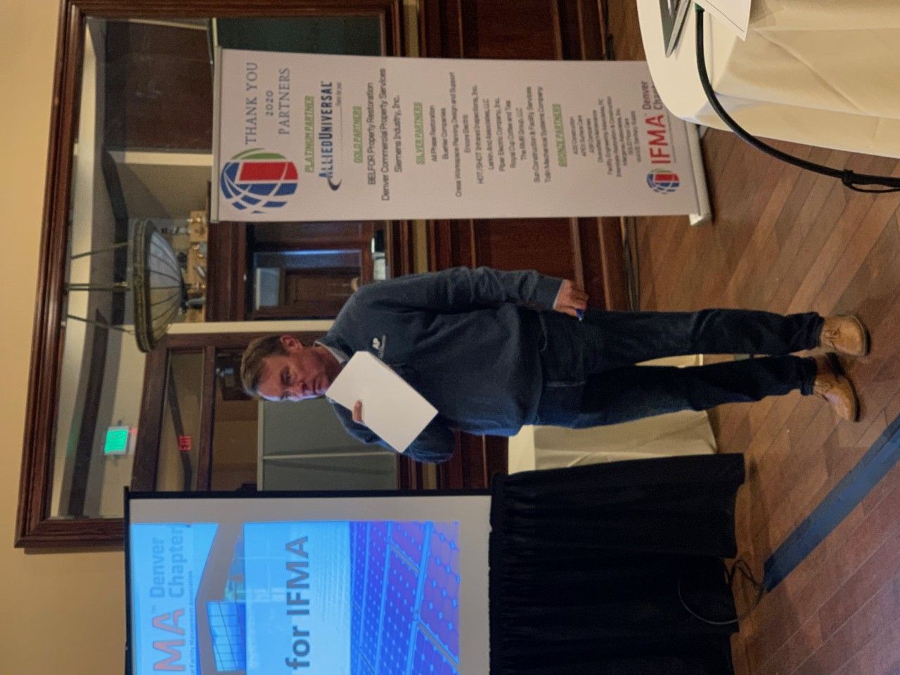 Man presenting at a conference, holding papers, with a banner and screen displaying text, in a room.