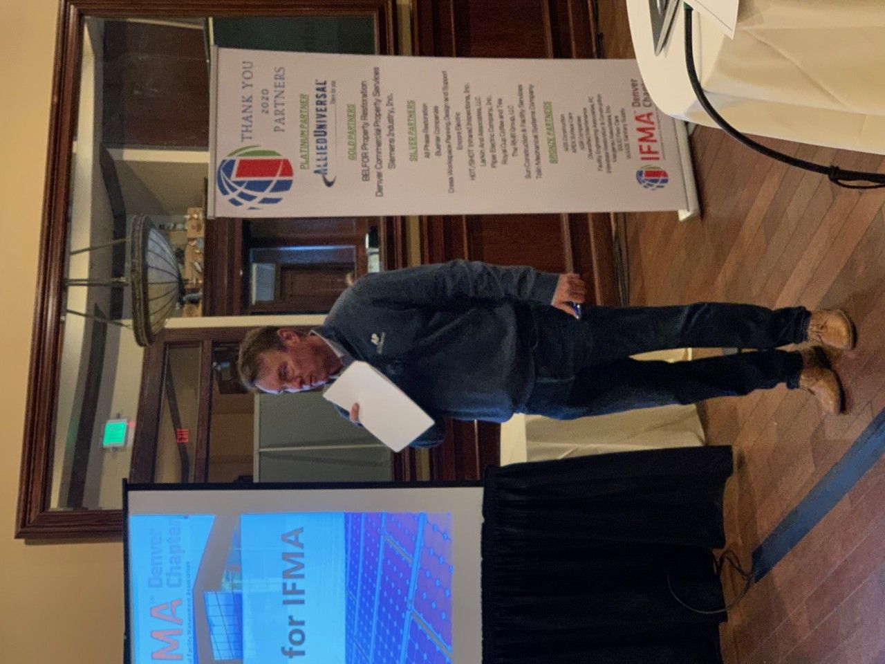 Man speaking at a conference. He stands near a screen and banner.