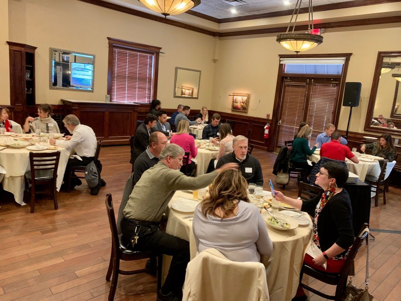 People seated at round tables in a large room, engaged in conversation. Dining. Wood floor, chandeliers.
