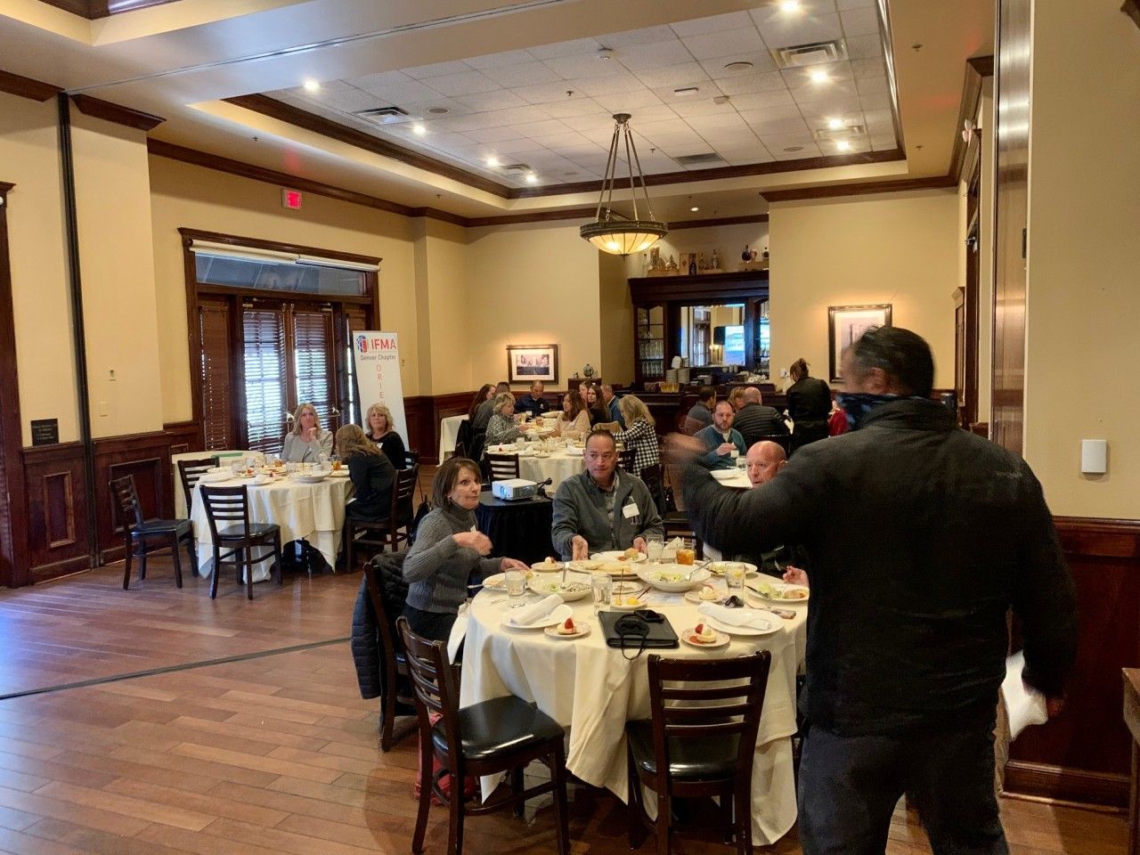 A man gestures while speaking in a restaurant. People sit at round tables. Dark wood trim with beige walls.