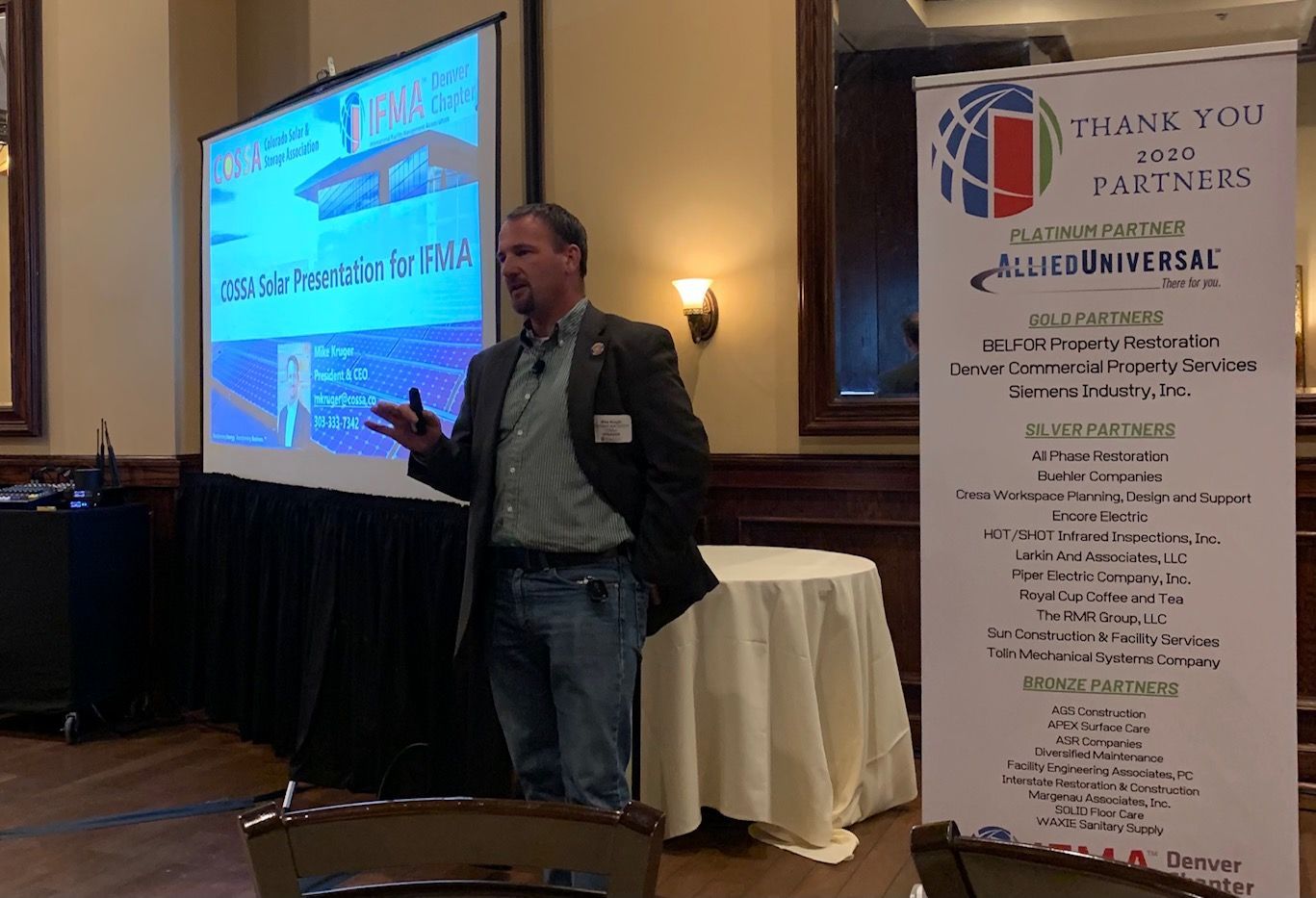 Man presenting at a conference, gesturing towards a projected slide, standing near a banner.