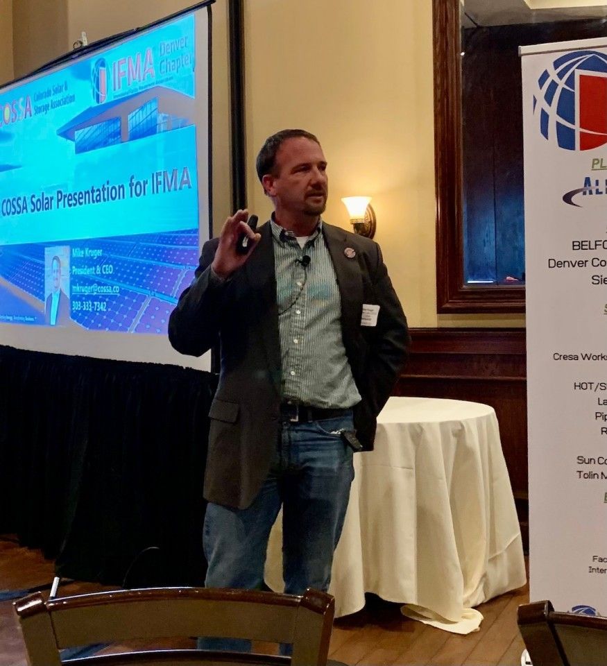 Man giving a presentation. He is wearing a blazer, jeans, and a name tag. A projector screen is behind him.
