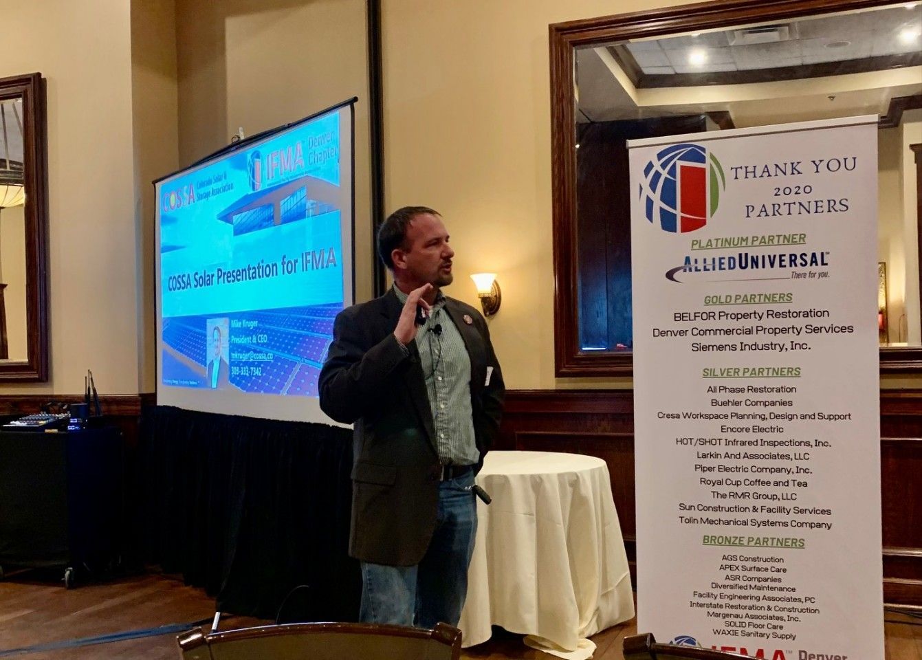 Man giving a presentation, standing near a screen displaying a diagram and a banner listing partners in a room.