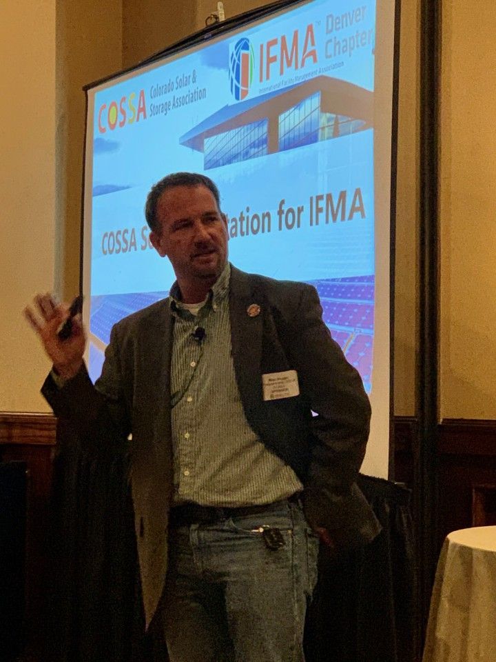 Man giving a presentation at a conference, gesturing with a hand near a projection screen.