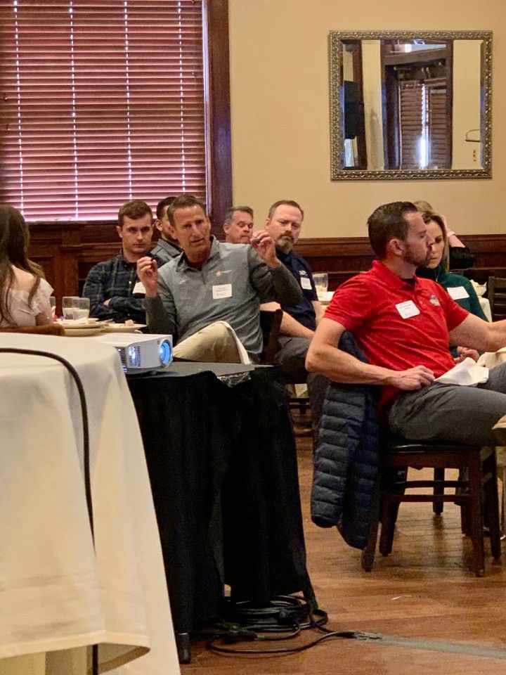 A man gestures while presenting to a seated audience at a meeting.