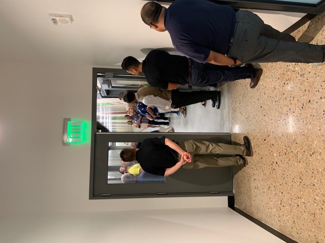 People standing in a hallway, looking toward an open doorway. The hallway has a beige floor and white walls.