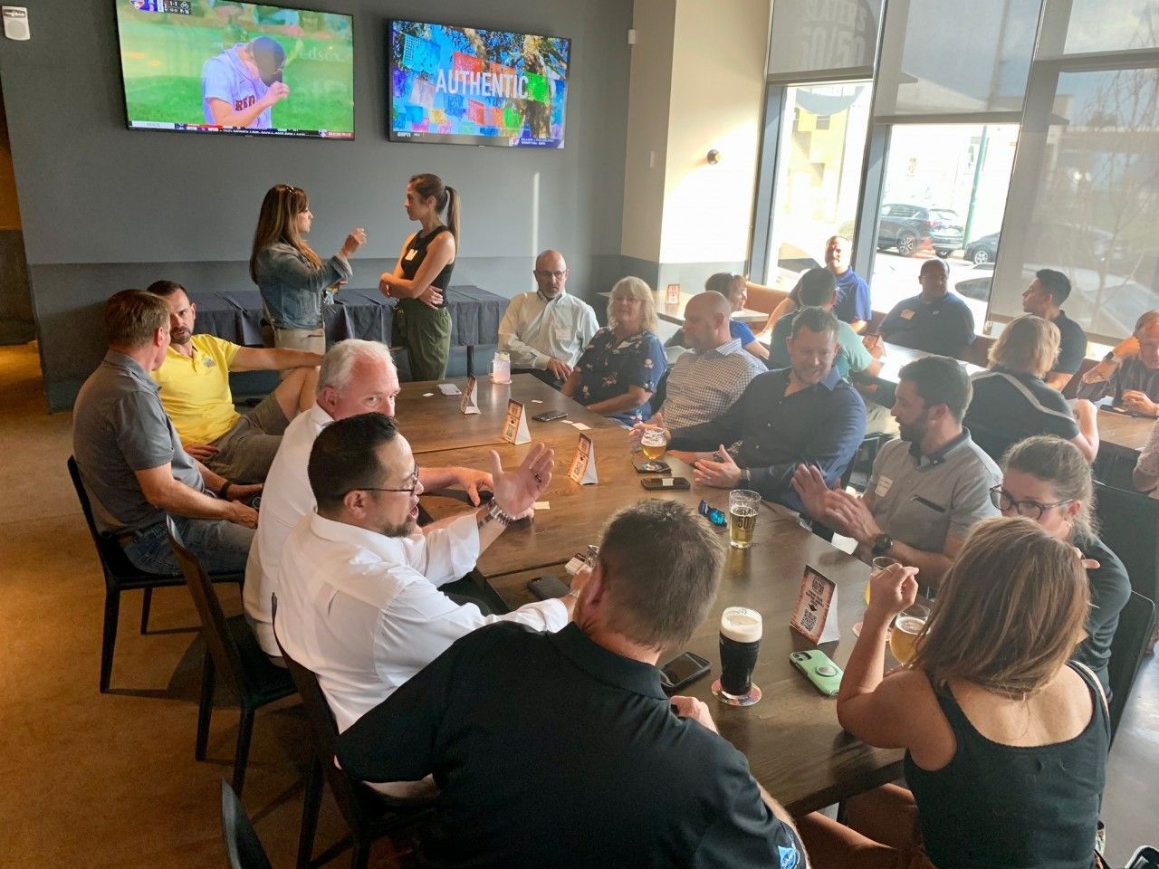 People gathered around a large table, two women standing, talking. Inside a restaurant, TVs on wall.