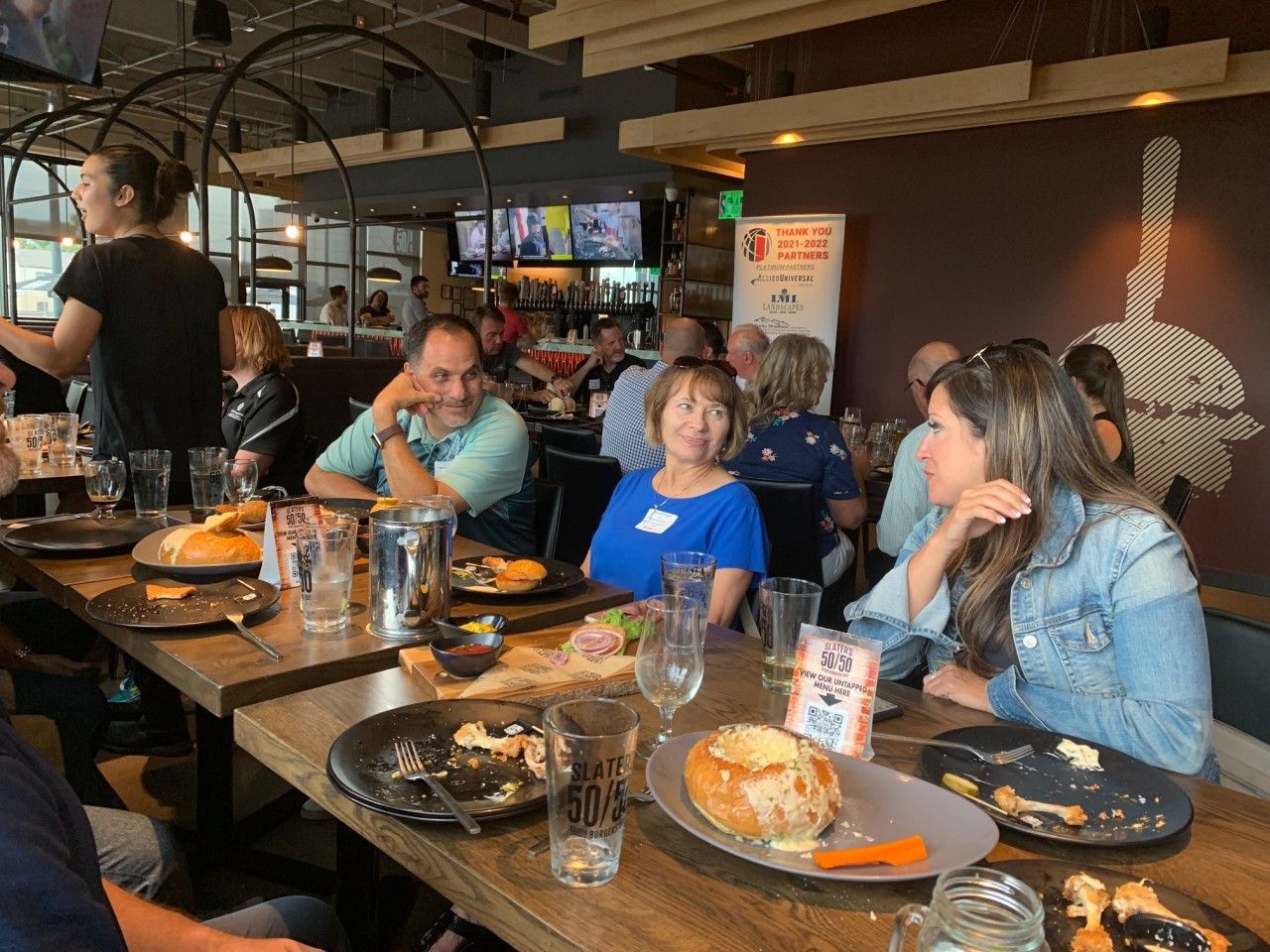 People seated around a long table in a restaurant; eating, conversing, and a server attending to the group.